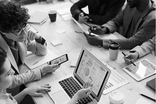 A close-up, black and white photo of professionals working at a desk. A laptop in the foreground displays detailed data dashboards and charts, while team members use smartphones and tablets to follow along.