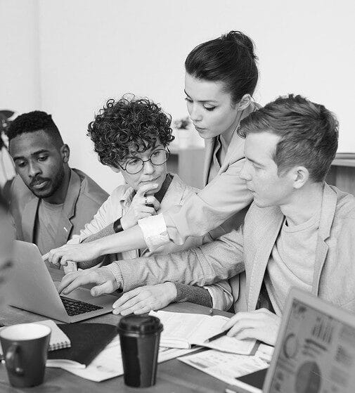 A black and white photo of four professionals huddled around a laptop in deep discussion, illustrating team collaboration and strategic planning.