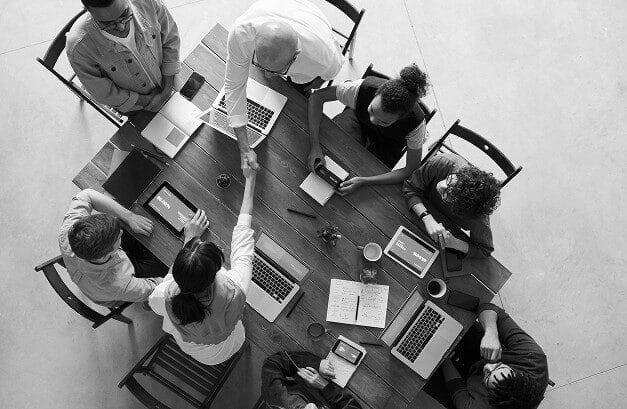 A black and white overhead view of eight people sitting around a large wooden table in a meeting. Two individuals are shaking hands over the center of the table, which is covered in laptops, tablets, and notebooks.